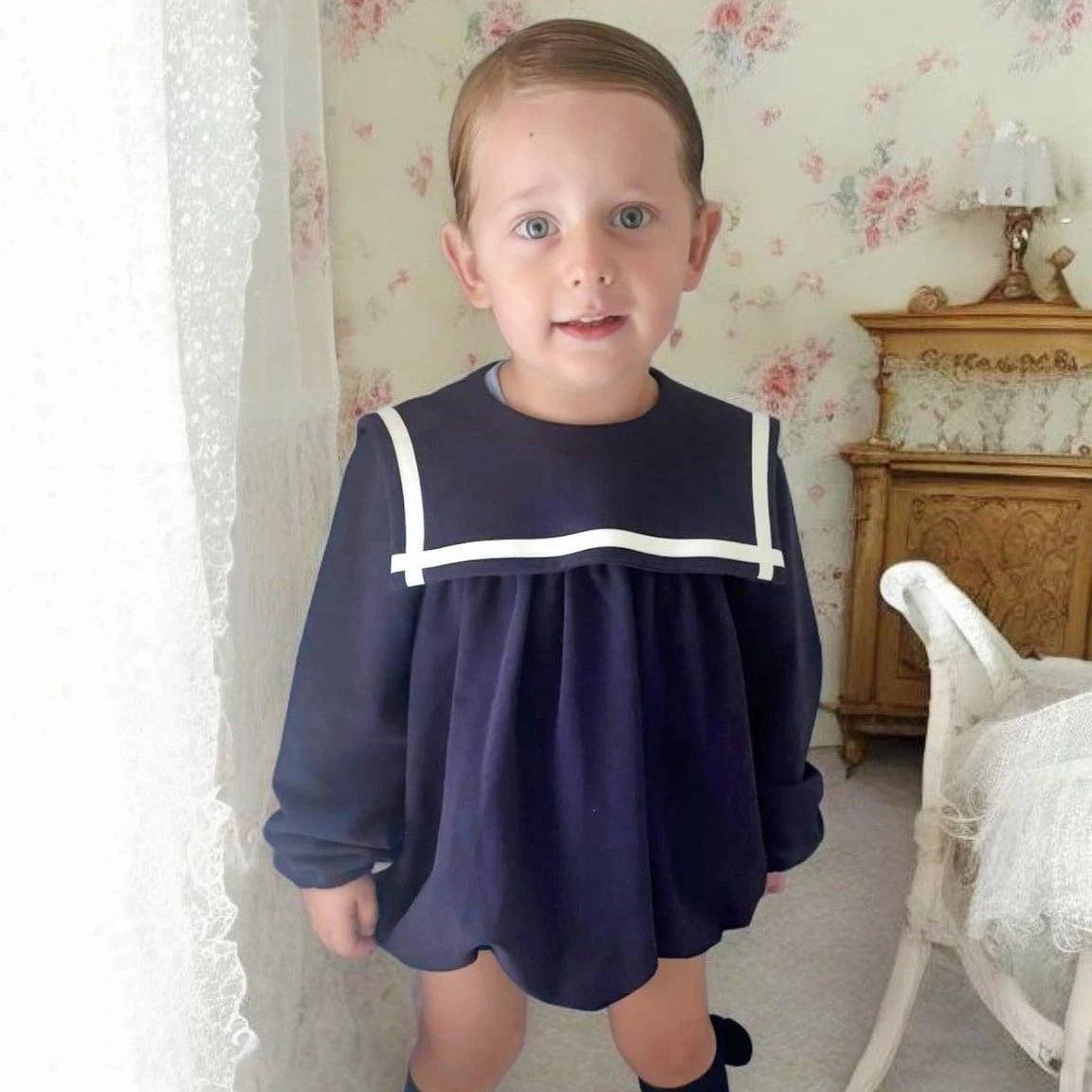 Child wearing a navy dress with white trim in a room with floral wallpaper.