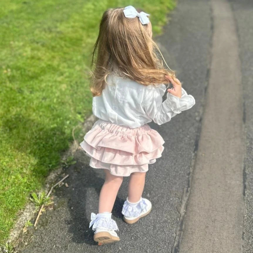 Child in a white blouse and pink skirt walking on a path with grass on one side.