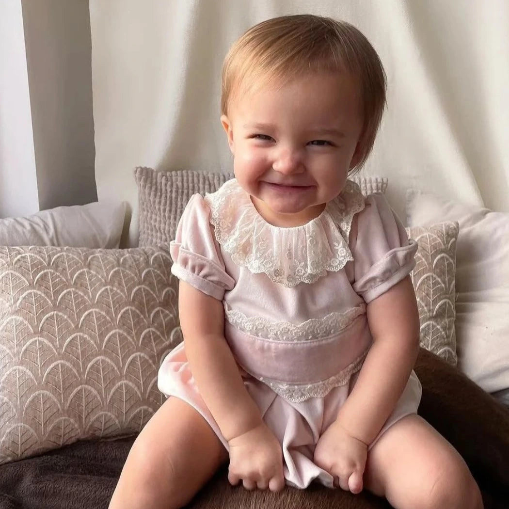 Child wearing a pink romper with lace details sitting on a couch.